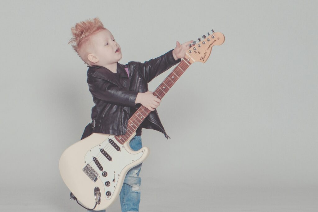 Stylish young boy with a mohawk strumming an electric guitar, wearing a leather jacket and blue jeans.